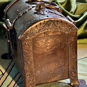 May include: A handcrafted copper chest with a domed lid and decorative metalwork. The chest features a handle, ornate detailing, and is supported by curved feet. The metal has a rich, aged patina, suggesting a vintage or antique aesthetic.
