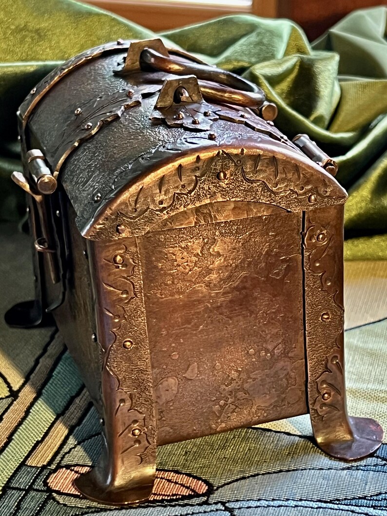 May include: A handcrafted copper chest with a domed lid and decorative metalwork. The chest features a handle, ornate detailing, and is supported by curved feet. The metal has a rich, aged patina, suggesting a vintage or antique aesthetic.