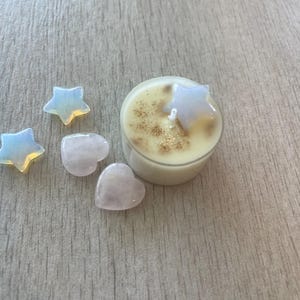 May include: A small, round candle with a star-shaped crystal on top, surrounded by star and heart-shaped crystals. The candle has a light-colored wax with gold glitter. The crystals are opalescent and pink.