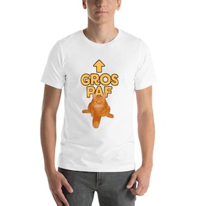 Può includere: T-shirt bianca con un gatto arancione dei cartoni animati e la scritta "GROS PAF" in giallo. Il gatto è seduto con le zampe divaricate e una freccia rivolta verso l'alto è sopra il testo. La maglietta è di stile casual.