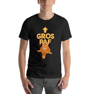Può includere: T-shirt nera con un gatto arancione dei cartoni animati e la scritta "GROS PAF" in giallo. Una freccia rivolta verso l'alto è sopra il testo. Il gatto è seduto con le gambe divaricate.