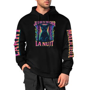 Może przedstawiać: Czarna bluza z kapturem z kolorową grafiką kota i napisem "JE VAIS TE TOUCHER LA NUIT". Rękawy mają słowa "LAKU" i "MALICIEUX" w kolorowej czcionce.