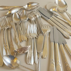 May include: A collection of vintage silverware, including spoons, forks, and knives. The silver utensils are arranged in a pile, reflecting light. The silverware has decorative detailing on the handles.