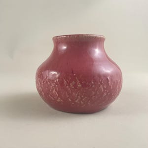 May include: A small, round, red-toned ceramic vase with a textured surface. The vase has a wide body and a narrow opening. The texture appears to be a pattern of small, irregular shapes. The rim of the vase is a lighter shade of red.