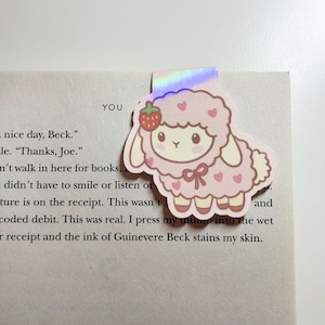 May include: A pink and white sheep bookmark with a strawberry on its head, placed on a book. The sheep has heart-shaped details and a bow. The bookmark is made of a holographic material.