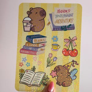 May include: A yellow sticker sheet with a glittery finish, featuring various illustrations. These include a brown cartoon animal with a drink, stacks of books, cherries, flowers, and the text "Books your greatest adventure."