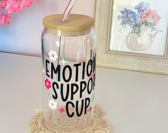 May include: A clear glass cup with a bamboo lid and a glass straw. The cup has the words "EMOTION SUPPORT CUP" in black, with floral and star accents. It sits on a beige macrame coaster. A framed floral print is in the background.