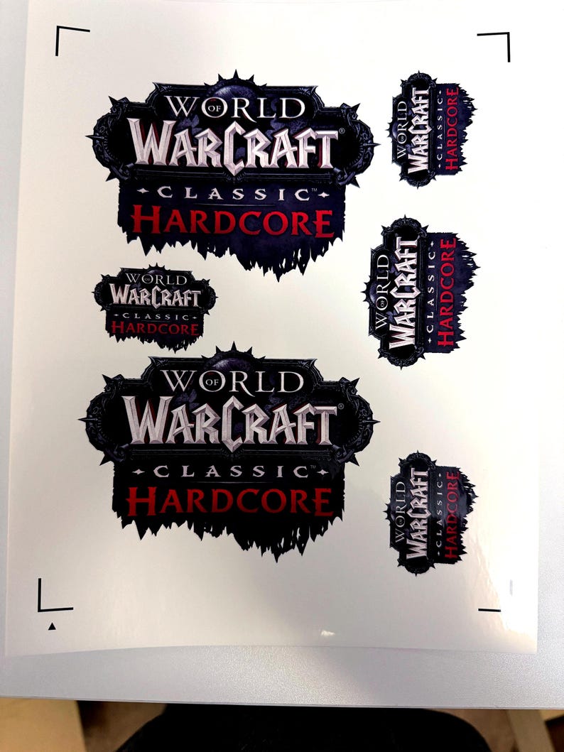 May include: A sheet of stickers featuring the "World of Warcraft Classic Hardcore" logo in various sizes. The logo is predominantly black, white, and red, with a stylized design. The stickers are printed on a white background.