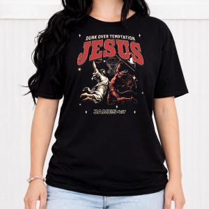 Könnte beinhalten: Schwarzes T-Shirt mit einer Grafik von Jesus, der einen Basketball gegen eine Teufelsfigur dunkt. Der Text "Dunk Over Temptation" und "JESUS" sind rot, darunter steht "JAMES 4:7". Das Design ist mittig auf dem Shirt platziert.