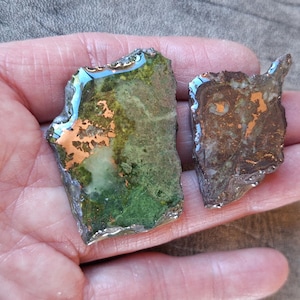 May include: Two irregularly shaped, decorative pieces with a metallic sheen. One is primarily green with copper accents, the other is brown with copper. Both have a silver-colored border and are held in a hand.