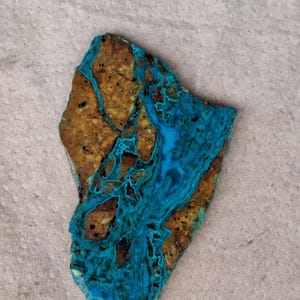 May include: A flat, irregularly shaped slab of chrysocolla stone featuring a vibrant, marbled pattern of bright turquoise blue veins interwoven with earthy, textured brown and tan mineral deposits across its polished surface.