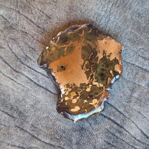 May include: A unique, irregularly shaped object with a metallic copper and green finish. The object has a glossy, reflective surface with hints of blue and is set against a textured, gray background.