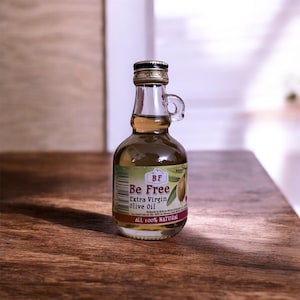 Può includere: Una piccola bottiglia di vetro di olio extra vergine di oliva "Be Free". La bottiglia ha un tappo dorato e un piccolo manico. L'etichetta presenta testo e un'illustrazione di ramo d'ulivo. L'olio è dorato e la bottiglia è su legno.