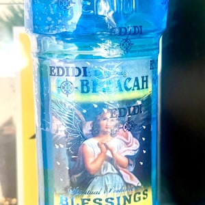 Puede incluir: Una botella de plástico transparente llena de líquido azul. La etiqueta presenta una ilustración de un ángel y las palabras "BLESSINGS" y "Spiritual Perfume". La botella tiene una tapa blanca con texto azul.
