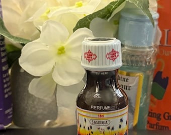 May include: A small perfume bottle with a white and red cap, labeled "PERFUME" and "SASORABIA." The bottle has a colorful label depicting a desert scene. Other perfume bottles and white flowers are in the background.