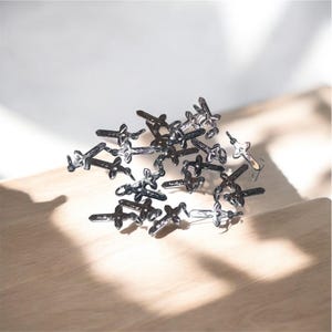 Può includere: Una collezione di piccoli ciondoli a forma di croce color argento. I ciondoli sono sparsi su una superficie di legno chiaro, con ombre che aggiungono profondità. Le croci hanno un design dettagliato e strutturato.