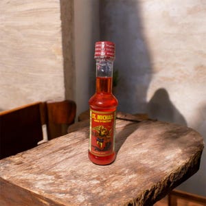 May include: A small glass bottle filled with a red liquid, labelled "ST. MICHAEL HUILE D'ONCTION". The bottle has a red cap and a label featuring an illustration of a saint. The bottle is on a wooden surface.