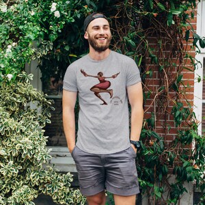 May include: A light gray t-shirt featuring a graphic of a dancing woman in a red dress with the text "THE UNIVERSAL MUSE WITH SARAH". The shirt is worn with gray shorts and a black cap.