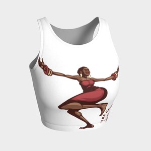 May include: White crop top featuring a stylized illustration of a woman in a red dress and jewelry, with arms outstretched. The design includes text at the bottom. The artwork has a vintage comic book style.