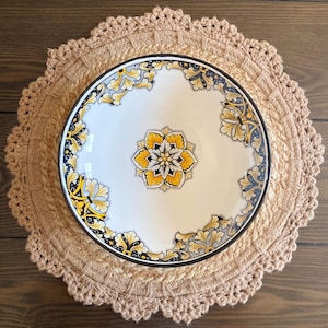 May include: A white ceramic bowl with a yellow and black floral design sits on a beige crocheted placemat. The bowl has a black rim and a central floral motif. The placemat is round with a scalloped edge, and the background is a wooden surface.