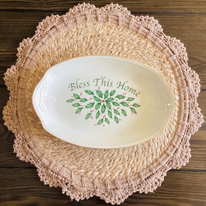 May include: Oval white serving platter with gold trim, featuring the text "Bless This Home" in green, surrounded by a holly and berry design. The platter rests on a round, beige crocheted placemat.