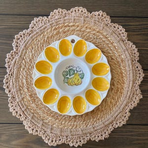 May include: White ceramic egg plate with 12 yellow egg-shaped indentations. The center features a yellow squash and green leaves design. The plate sits on a round, woven placemat.