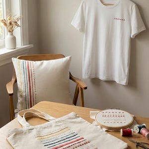Peut inclure: Ensemble d'articles brodés à la main, dont un t-shirt blanc, un coussin et un sac fourre-tout. Les articles présentent des motifs brodés colorés. Un cercle à broder avec un motif et des bobines de fil sont également visibles.