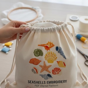 Peut inclure: Un sac à cordon de couleur crème avec une broderie de coquillages colorés. Le sac porte le texte "SEASHELLS EMBROIDERY PDF Digital Pattern". Le sac est tenu par une personne.