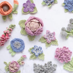 Puede incluir: Una colección de flores de ganchillo hechas a mano en varios diseños. Las flores incluyen flores naranjas, moradas, rosas, azules y grises con hojas verdes. La disposición está sobre una superficie blanca, mostrando la artesanía detallada.