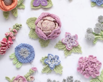 May include: A collection of colorful, handmade crochet flowers in various designs. The flowers include orange, purple, pink, blue, and gray blooms with green leaves. The arrangement is on a white surface, showcasing the detailed craftsmanship.