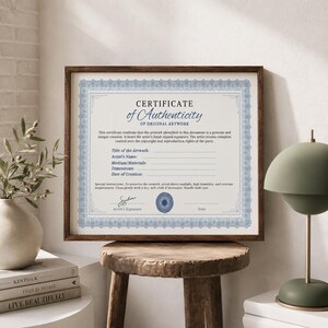 Puede incluir: Un certificado de autenticidad enmarcado con un borde azul y blanco. El certificado incluye campos de texto para el t&iacute;tulo de la obra de arte, el nombre del artista y las dimensiones. El marco es marr&oacute;n y el certificado se muestra en un taburete de madera.