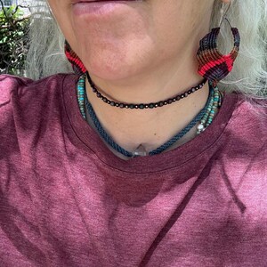 May include: A pair of teardrop-shaped macrame earrings featuring a chevron pattern in shades of black, brown, and red. The jewelry set includes a dark beaded choker, a blue braided cord necklace with a clear crystal pendant, and turquoise beads.