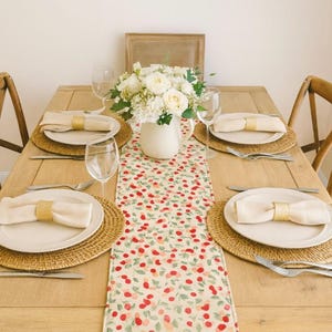 May include: A dining table set for a meal, featuring a floral table runner with red and green accents. Each place setting includes a woven placemat, white plate, folded napkin with a gold ring, and silverware. A white vase holds a bouquet of white flowers.
