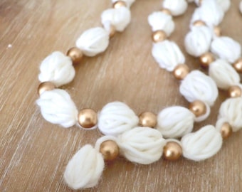 May include: A decorative garland featuring alternating white yarn pom-poms and smooth metallic gold beads strung together. The white yarn elements are textured and soft, while the gold beads provide a reflective, polished contrast throughout.