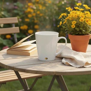 May include: A white ceramic mug with a small bee illustration near the base, featuring a curved handle. The mug is positioned on a round wooden table alongside an open book, a folded beige cloth napkin, and a terracotta pot with yellow flowers.