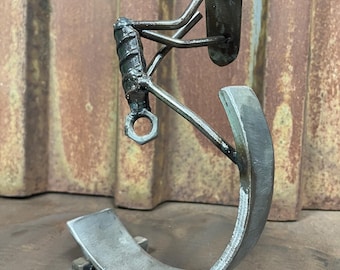 May include: A handcrafted metal sculpture featuring a curved base and an abstract figure. The sculpture is made of welded steel with a textured finish. The figure has a head, arms, and a torso. The sculpture is approximately 10 inches tall.
