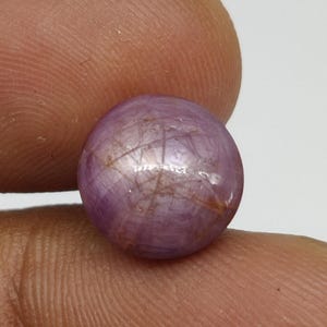 May include: A spherical, polished, purplish-pink gemstone. The gem exhibits a star-like effect, with lines emanating from the centre. The stone is held between two fingers, against a plain white backdrop.