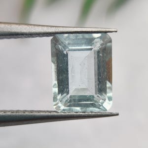 Puede incluir: Una gema rectangular de color azul claro es sostenida por unas pinzas plateadas. La gema es transparente con múltiples facetas. La gema es de color azul claro. Las pinzas son de metal.