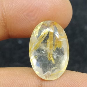 以下が含まれることがあります： 透明感のある、金色がかった黄色のオーバルカットの宝石。石には金色の針状の内包物があり、独特の視覚的テクスチャを生成しています。暗い背景を背景に、指で宝石が持たれています。