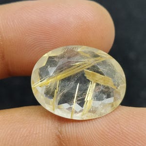以下が含まれることがあります： 透明感のある、金色の輝きを持つ楕円形のファセット宝石。内部には金色の針状のインクルージョンがあり、独特の模様を作り出しています。暗い背景の中で、指で宝石が持たれています。