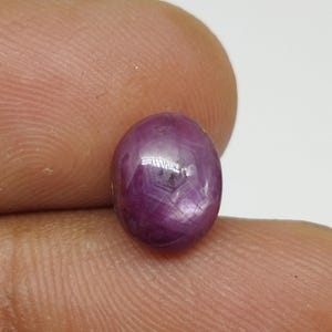 May include: A polished, oval-shaped, deep red gemstone. The gem has a star-like pattern visible on its surface. The stone is held between two fingers, showcasing its size and colour. This gemstone is likely a ruby.