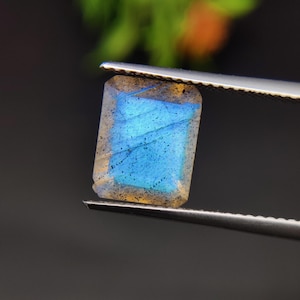 May include: A rectangular, polished gemstone with a vibrant blue and yellow iridescence, held by silver tweezers. The stone's surface displays a play of light, with a mix of blue and yellow hues. The background is blurred, highlighting the gem's details.