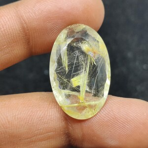 以下が含まれることがあります： 透明感のある黄色がかった楕円形の宝石。内部には金色の針状のインクルージョンが見られます。宝石はファセットカットされており、光を反射します。全体的に半透明で、表面は研磨されています。指で宝石を持っています。