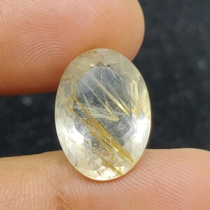 以下が含まれることがあります： 透明感のある、金色のイエローカラーのオーバルカットの宝石。内部には金色の針状のインクルージョンがあり、独特の模様を作り出しています。暗い背景を背景に、指で宝石が持たれています。