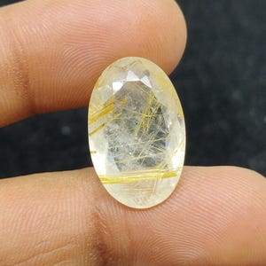 以下が含まれることがあります： 透明感のある、金色のイエローカラーのオーバルカットの宝石。繊細な金色の針状のインクルージョンが全体にあり、独特の模様を作り出しています。指で宝石を持ち、磨かれた表面と内部のディテールを際立たせています。