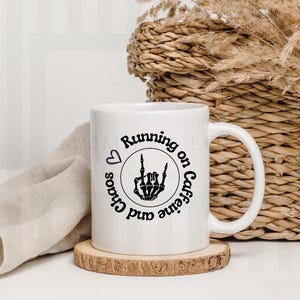 May include: White ceramic mug with a black graphic that reads "Running on Caffeine and Chaos" with a heart and a skeleton hand. The mug sits on a small wooden coaster. A woven basket and dried plants are in the background.
