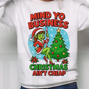 May include: White sweatshirt with a graphic featuring a green elf in a Santa hat, holding a candy cane next to a Christmas tree. The text reads "MIND YO BUSINESS" and "CHRISTMAS AIN'T CHEAP" in red and green.
