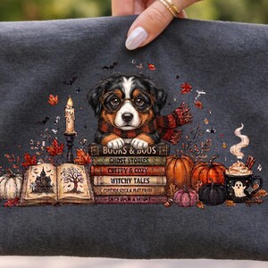 May include: A dark gray t-shirt featuring a graphic of a puppy wearing glasses and a scarf, resting on a stack of books titled Books & Boos, Ghost Stories, Creepy & Cozy, Witchy Tales, Pumpkin Spice & Plot Twist, and Once Upon a Fright.