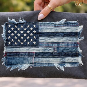 May include: A dark grey fabric featuring a denim patch of the American flag. The flag is constructed from various shades of blue denim, with white embroidered stars and red stitching. The edges of the flag are frayed.