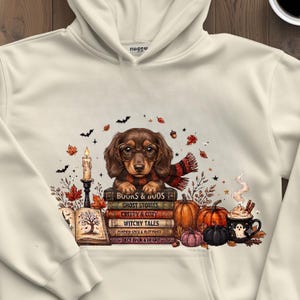 Può includere: Una felpa con cappuccio color crema con la grafica di un bassotto marrone che indossa occhiali e una sciarpa, appoggiato su una pila di libri intitolati Books & Boos, Ghost Stories, Creepy & Cozy, Witchy Tales, Pumpkin Spice & Plot Twist e Once Upon a Fright.